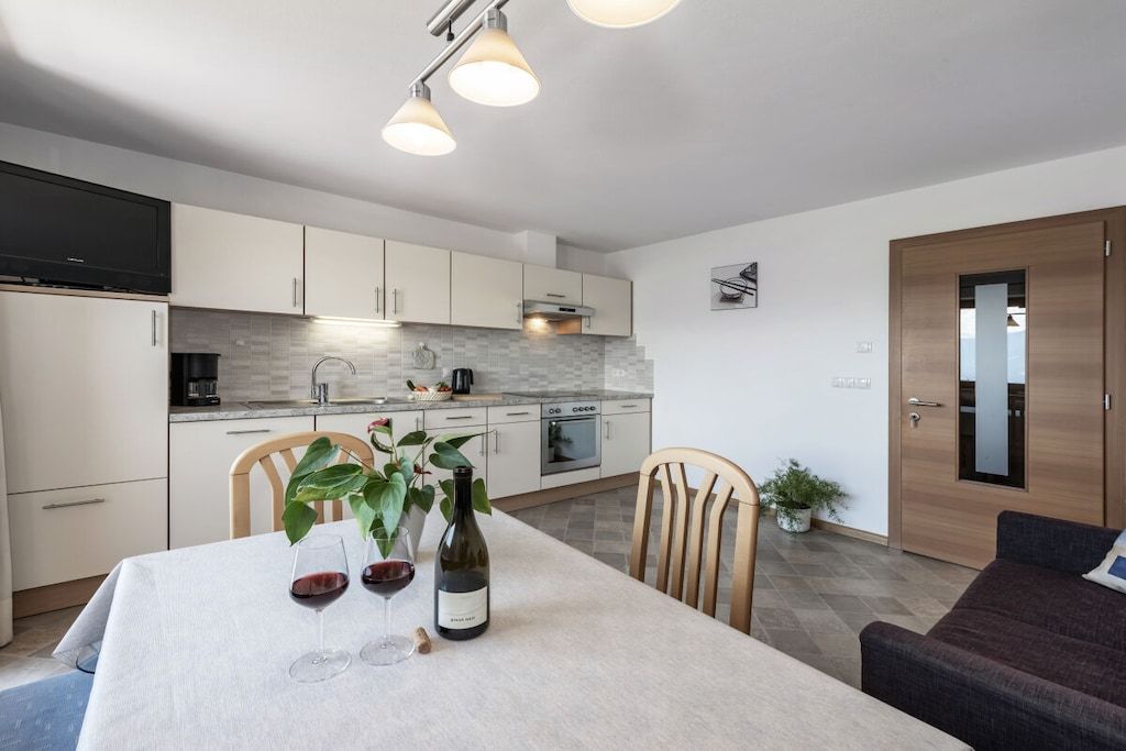 A modern kitchen with white cabinets, a dining table with a wine bottle and glasses, and a sofa in the adjacent living area.