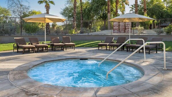 A hot tub surrounded by lounge chairs and umbrellas, with a pool in the background.