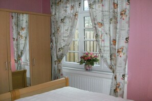Un dormitorio con una cama de madera, un armario, un ventanal con cortinas florales y un florero con flores.