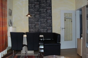 A room with a dining table and three chairs, a radiator, a wall with a coffee shop pattern, and a door with a curtain. - Holiday apartment with 1 bedroom (Borkum)
