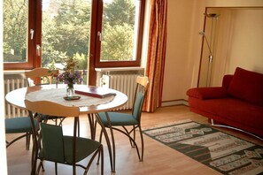 A room with a round table, chairs, a red sofa, and a view of trees through large windows. - Villa Renate on the North Sea (Cuxhaven)