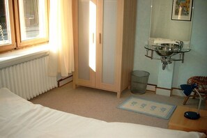 Een slaapkamer met een bed, een radiator, een kledingkast, een badkamer met een glazen wastafel en een raam met gordijnen.