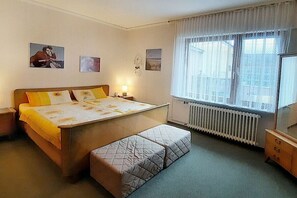 Une chambre à coucher avec un lit, une table de chevet, une lampe, une fenêtre avec des rideaux et un radiateur.