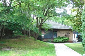 Une maison de plain-pied, dotée d’un toit en pente, entourée d’une végétation luxuriante et d’arbres.