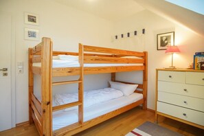 Un lit superposé avec un matelas blanc et une structure en bois, une commode blanche avec des tiroirs et une lampe posée sur la commode.