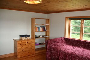 A room with a wooden dresser, a bookshelf, a patterned sofa, and a window with a view of greenery.