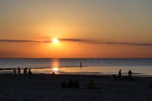 A beach scene with people sitting and standing on the sand, a pier extending into the water, and the sun setting in the background. - ||--------------------House at Domburg----------------|| (Domburg)