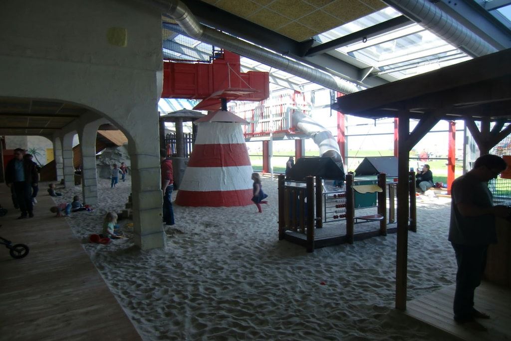 Un espacio interior con suelo de arena, una estructura grande de color rojo y blanco, y una zona de estar cubierta.