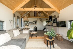 Ein modernes Wohnzimmer mit hoher Decke, Holzbalken, einem grauen Sofa, einem Essbereich und einem an der Wand montierten Fernseher.