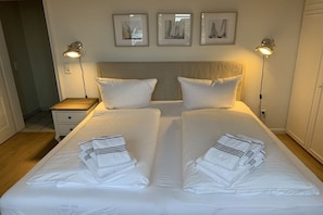 Cama doble con sábanas y toallas blancas, lámparas de noche y cuadros en la pared.