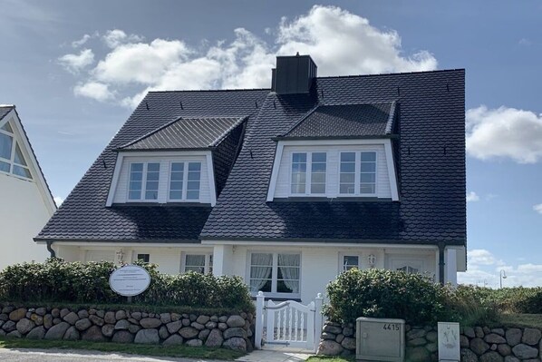 Una casa de dos pisos con techo oscuro, ventanas blancas y un muro de piedra con una reja.