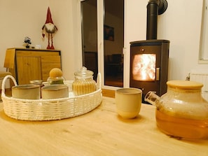 A room with a wood-burning stove, a wicker basket with jars, a mug, and a teapot on a wooden table.