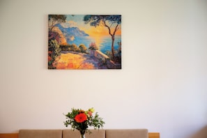 A painting of a landscape with trees and a pathway, a vase with flowers on a table, and a beige sofa.