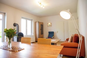 A modern living room with a wooden dining table, a blue armchair, and a sofa. There is a radiator, a window, and a picture on the wall.