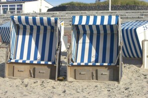 Des chaises de plage avec des parasols à rayures, une plage de sable et un bâtiment avec une enseigne visible à l’arrière-plan.