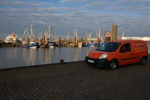 Ein roter Van ist in der Nähe eines Uferbereichs geparkt, im Hintergrund liegen Boote im Hafen.