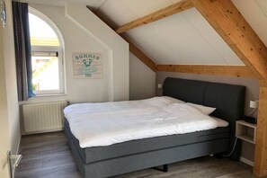 Quarto com teto inclinado, cama com roupa de cama branca, mesa de cabeceira pequena e luminária fixada na parede.
