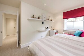 A bedroom with a bed, a desk, a window with a red valance, and a door leading to another room. - Georgian Elegance in Islington, by Veeve (London)