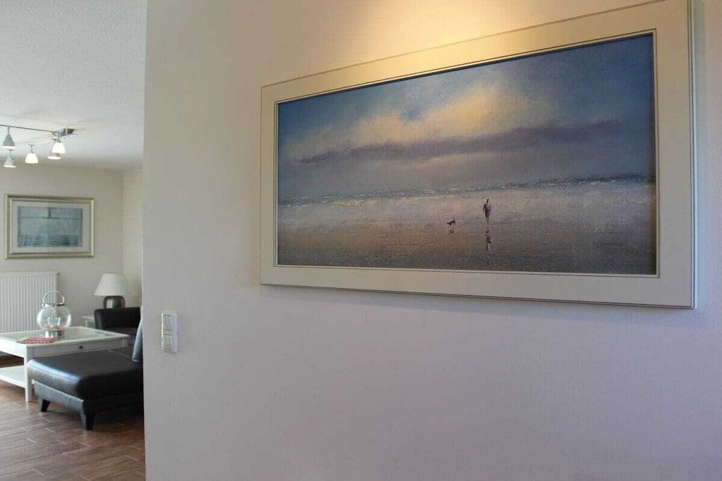 A room with a large framed painting of a beach scene, a small table with a lamp, and a chair.