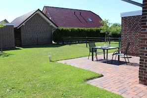 A brick house with a red roof, a paved patio with a table and chairs, and a well-maintained lawn. - Holiday home with 2 bedrooms (Greetsiel)