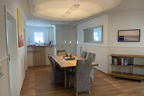 A dining area with a wooden table and chairs, a kitchen in the background, and a bookshelf with books. - Holiday apartment with 2 bedrooms (Hooksiel)
