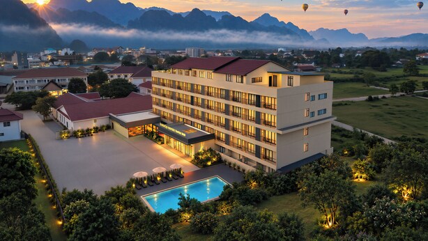 A hotel with multiple floors, a swimming pool, and a view of mountains and hot air balloons in the distance. - Vangvieng THAVIKUN HOTEL (Vang Vieng)