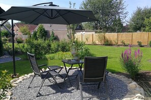 Une terrasse extérieure avec une table et des chaises, un grand parasol, et un jardin agrémenté d’une clôture en bois et de diverses plantes.
