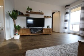 A living room with a flat-screen TV, wooden entertainment unit, potted plants, and a large window. - Holiday apartment with 1 bedroom (Rhauderfehn)