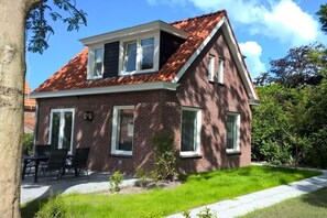 Ein zweistöckiges Backsteinhhaus mit rotem Ziegeldach, einer Veranda mit Sitzgelegenheiten und einem kleinen Garten.