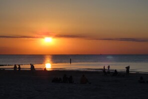 Eine Strandansicht mit Menschen, die auf dem Sand sitzen und stehen, und einem Sonnenuntergang über dem Wasser.