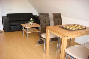 Ein Zimmer mit einer schwarzen Couch, einem kleinen Holztisch, zwei Korbstühlen und einem Esstisch aus Holz.