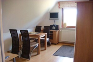 Ein kleiner, möblierter Raum mit Esstisch, Stühlen, einem Fernseher und einem Fenster mit Vorhängen.