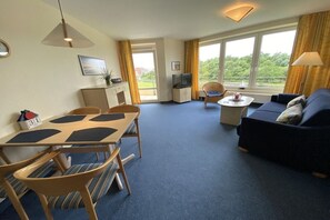 A hotel room with a dining table, chairs, a sofa, a TV, and a balcony with curtains. - 120 Kurpark Residenz Cuxhaven (Cuxhaven)