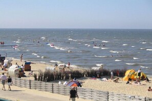 Ein Strand mit badenden und am Ufer entlang gehende Menschen, Zelten und einem Holzzaun.
