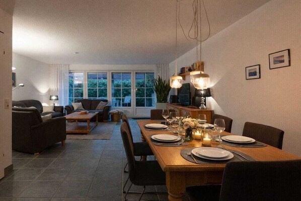 Ein Essbereich mit einem Holztisch, gedeckt für vier Personen, umgeben von Stühlen. Im Hintergrund befindet sich eine Couch und ein kleiner Tisch mit einer Lampe.