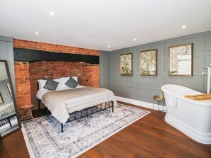 A bedroom with a brick wall, a bed with a white duvet, a patterned rug, a white bathtub, and framed artwork on the wall. - The Annex at The Lodge (Shrewsbury)