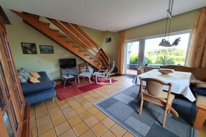 Ein Wohnzimmer mit Treppe, Fernseher, Essbereich mit Tisch und Stühlen sowie einer Couch.