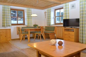 Ein gemütliches Zimmer mit Holzbalkendecke, einem Esstisch mit Stühlen, einem Fernseher und einem Kreuz an der Wand.