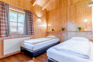 Ein Zimmer mit zwei Betten, Holzwänden, einem Fenster mit Vorhängen, einem Heizkörper und einem Regal mit Pflanzen.