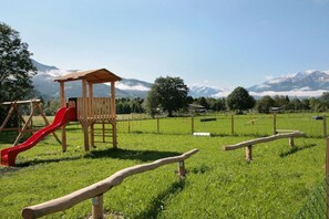 Ein Spielplatz mit einer Holzspielkonstruktion und einer roten Rutsche, vor einer Bergkulisse und blauem Himmel.