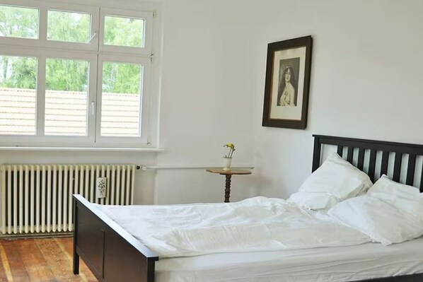 A bedroom with a bed, a window, a radiator, and a small table with a vase.