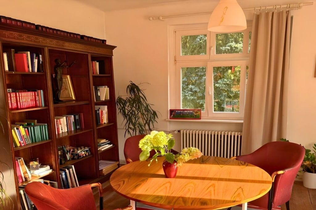Une pièce avec une bibliothèque en bois, une table ronde ornée d’un vase de fleurs et une fenêtre avec des rideaux.