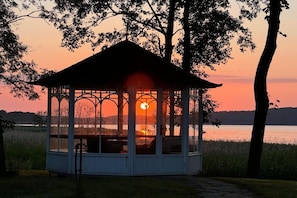 Ein Pavillon mit Dach und offenen Seiten, in Wassennähe mit Sonnenuntergang im Hintergrund.