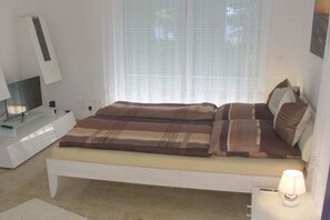 Een slaapkamer met een bed, nachtkastje met lamp, televisie en raam met zonwering.