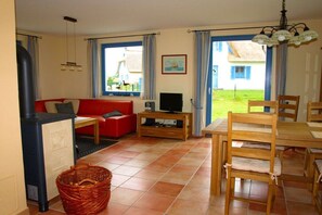 Ein Wohnzimmer mit einem roten Sofa, einem Fernseher auf einem Holzgestell, einem Esstisch mit Stühlen und einem großen Fenster mit Vorhängen.
