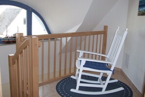 Ein weißer Schaukelstuhl mit blauer Kissenauflage auf einem dunklen, runden Teppich, platziert in der Nähe einer Holztreppe mit weißer Geländer.