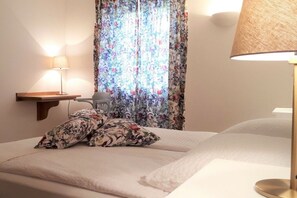 A bedroom with a bed, two pillows, a bedside table with a lamp, and a window with floral curtains. - Holiday home Swallow Heaven (Poseritz\/Rügen OT Mellnitz - Hof)