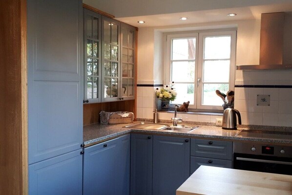 A kitchen with blue cabinets, a window with white curtains, a sink, and a kettle. - Holiday home Swallow Heaven (Poseritz\/Rügen OT Mellnitz - Hof)