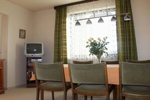 Ein Essbereich mit Tisch und Stühlen, einem Fernseher auf einem Schrank und einem Fenster mit Vorhängen.