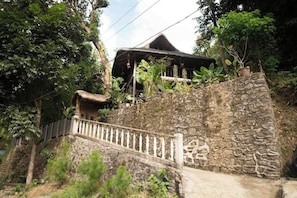 Eine Steintreppe führt zu einem Haus mit einem Strohdach, umgeben von üppiger Vegetation.
